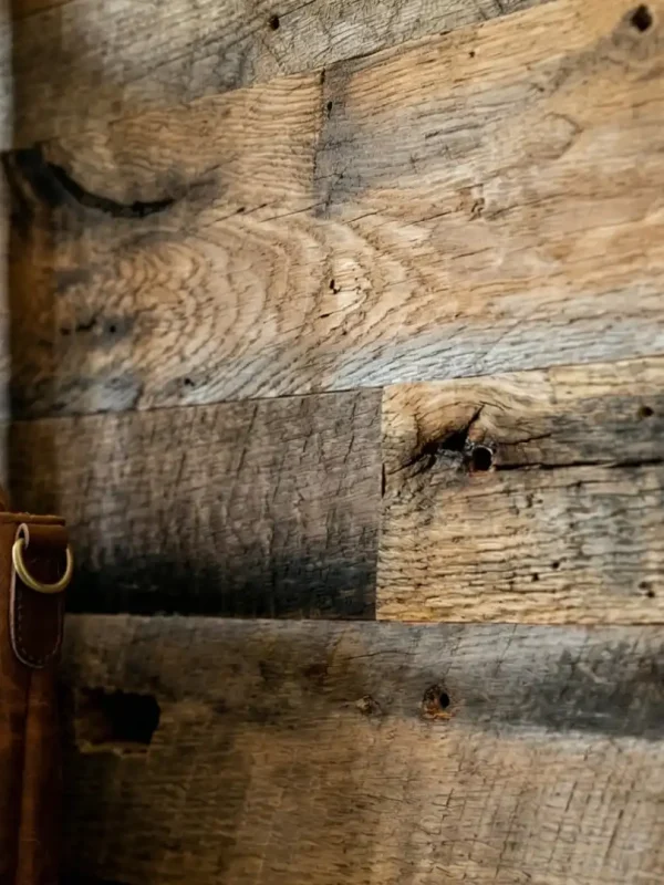Close-up view of a brown leather tote bag hanging from a turned wooden peg, showcasing a detailed contrast against an accent wall of distressed, reclaimed barnwood planks. The warm lighting highlights the natural grain variations and original saw marks of the weathered brown barn wood paneling.