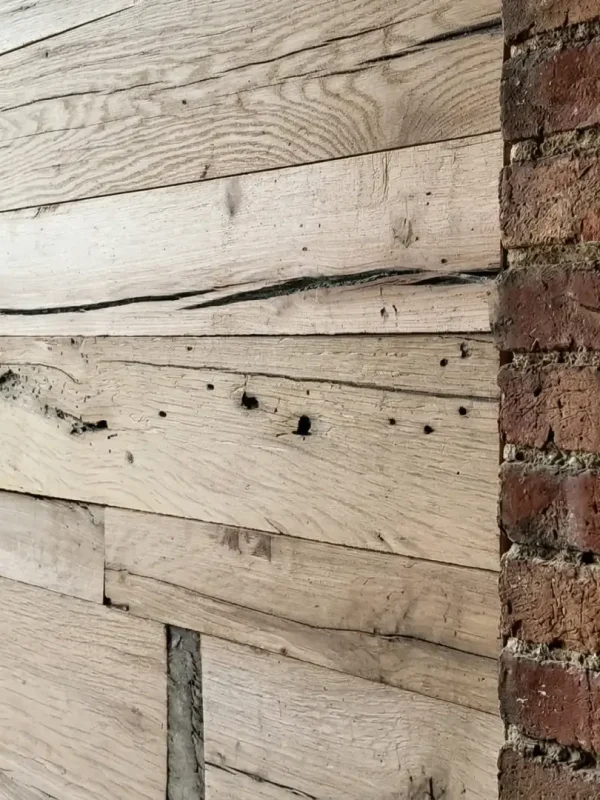 Architectural close-up showing the flush transition between horizontal reclaimed oak cladding and rough exposed brick. The detail emphasizes the precision milling, tight seams, and natural checking of the beam-sawn timber.